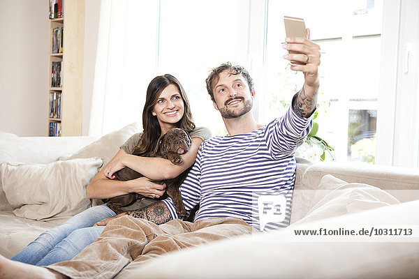 Paar sitzend mit Olde English Bulldogge auf der Couch mit einem Selfie mit Smartphone