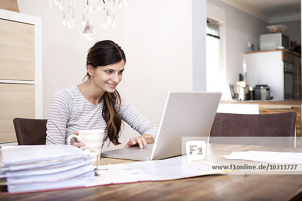 Lächelnde Frau am Holztisch sitzend mit Tasse Kaffee und Laptop