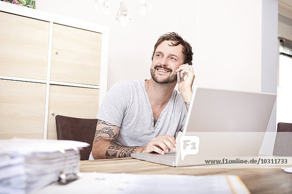Smiling man sitting at wooden table with laptop and folder telephoning with smartphone