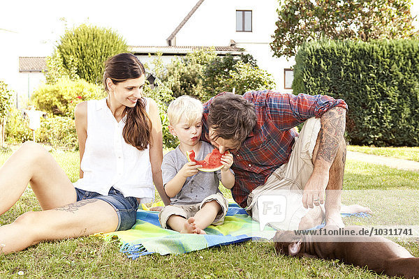 Eltern sitzen mit ihrem kleinen Sohn auf einer Decke im Garten und essen Wassermelone.