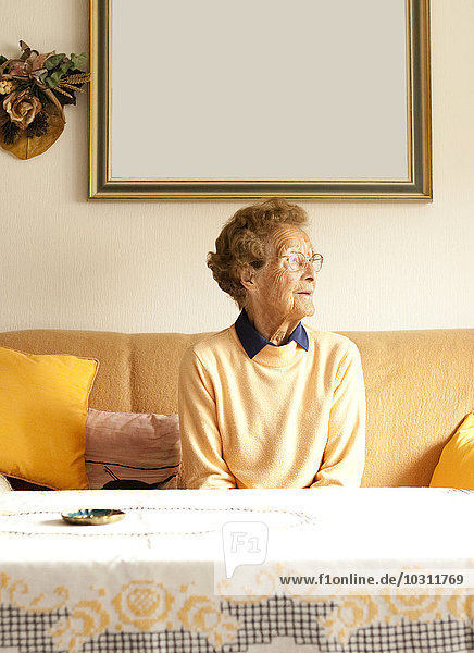 Porträt einer alten Frau  die auf der Couch in ihrem Wohnzimmer sitzt.