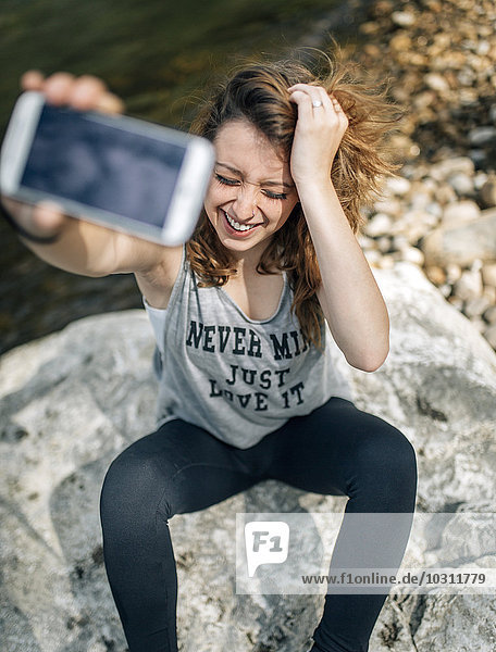 Porträt einer lachenden jungen Frau,  die auf einem Felsen sitzt und sich selbst mit einem Smartphone nimmt.