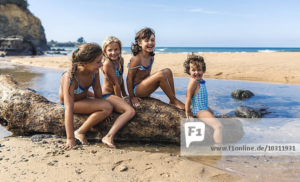 Spain  Colunga  four girls sitting on dead wood on the beach
