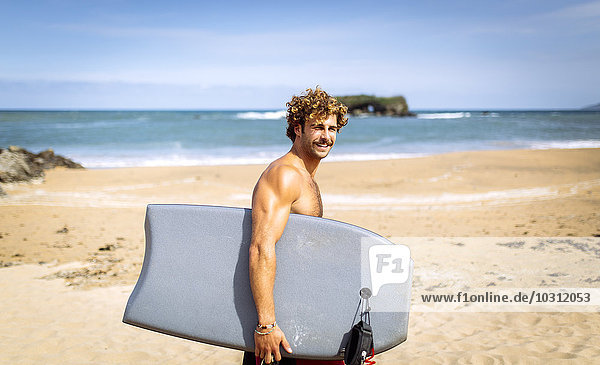 Spanien  Asturien  Colunga  lächelnder Surfer beim Wandern mit Bodyboard am Strand