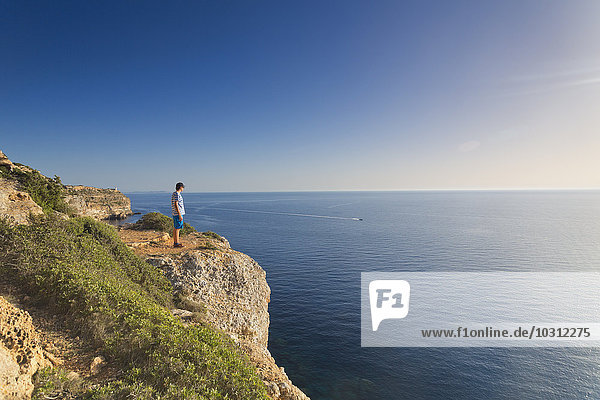 Spain  Balearic Islands  Majorca  one teenage boy standing on a rock at the cliff coast  watching the sundown