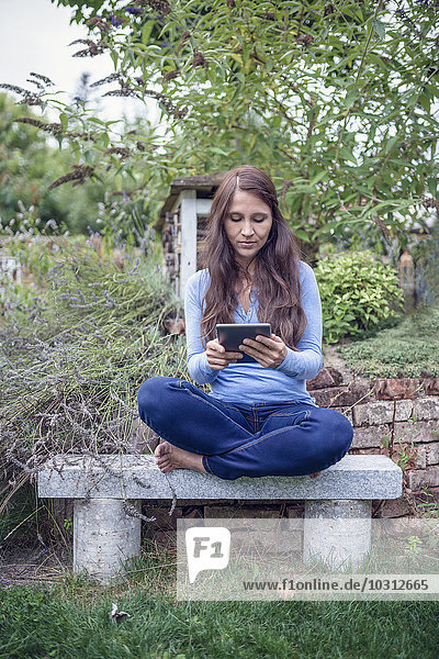Woman sitting cross-legged on a bench in the garden looking at digital tablet
