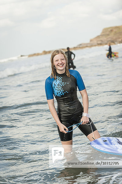 Porträt eines lächelnden Teenagermädchens mit Surfbrett im Neoprenanzug