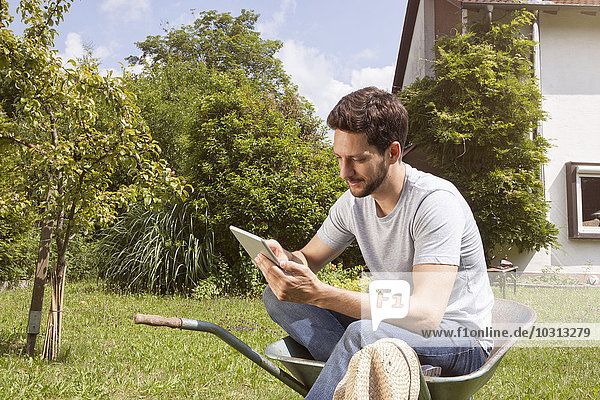 Mann sitzend in Schubkarre im Garten mit digitalem Tablett