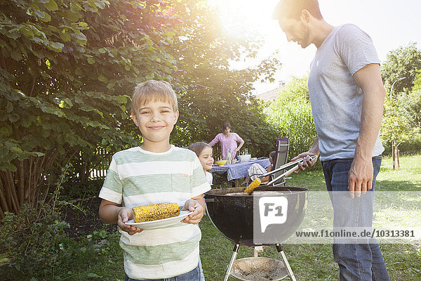 Lächelnder Junge mit Maiskolben auf einem Familiengrill im Garten