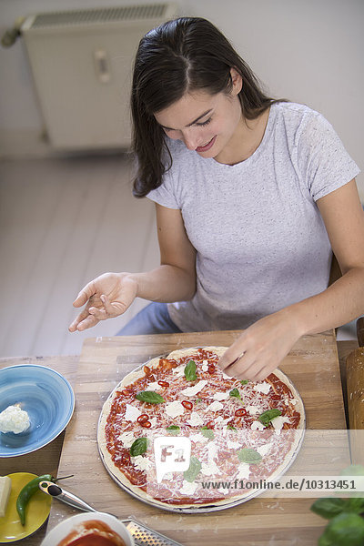 Junge Frau bereitet Pizza mit Mozzarella  Chilischoten und Basilikum zu.