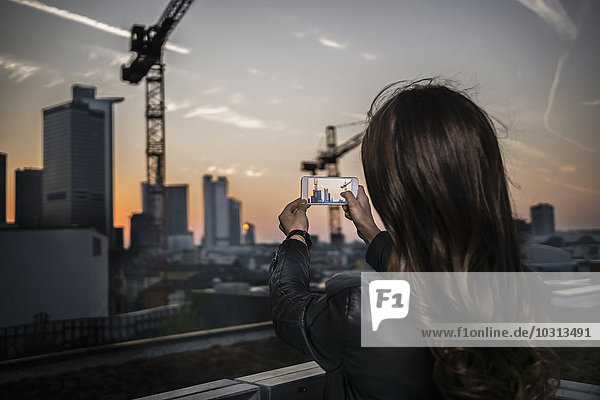 Deutschland  Frankfurt  Rückansicht einer jungen Frau  die in der Dämmerung mit dem Smartphone fotografiert.