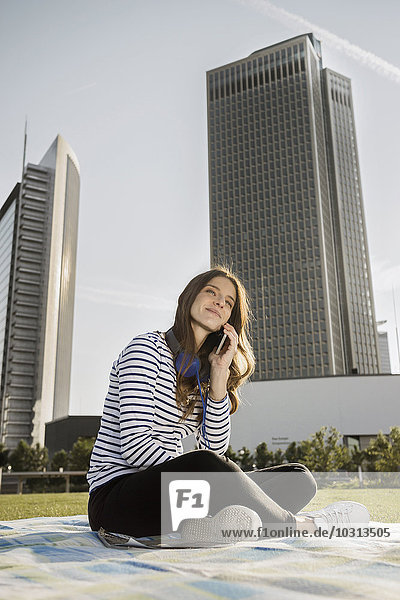 Deutschland  Frankfurt  junge Frau auf einer Wiese sitzend mit Smartphone telefonierend