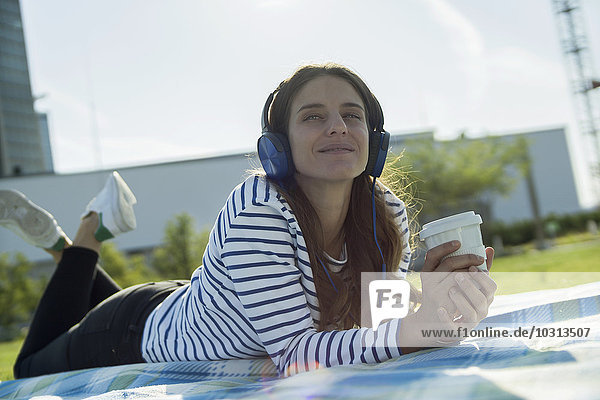 Junge Frau mit Kaffee zum Liegen auf einer Wiese und Musik hören mit Kopfhörer