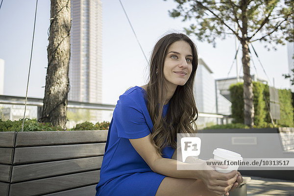 Deutschland  Frankfurt  Porträt einer lächelnden Geschäftsfrau auf einer Bank mit Kaffee zum Mitnehmen