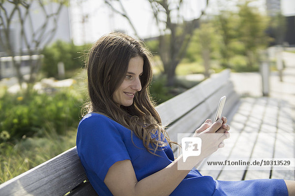 Lächelnde Geschäftsfrau sitzt auf einer Bank und schaut auf das Smartphone.