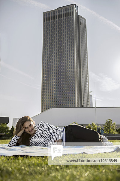 Deutschland  Frankfurt  Porträt einer lächelnden Frau  die auf einer Wiese vor dem Wolkenkratzer liegt.