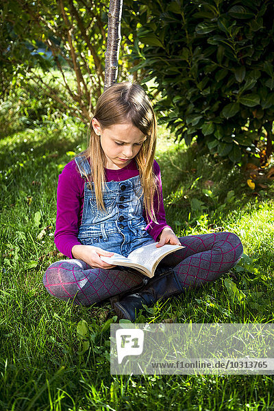 Little girl sitting on a meadow in the garden reading a book