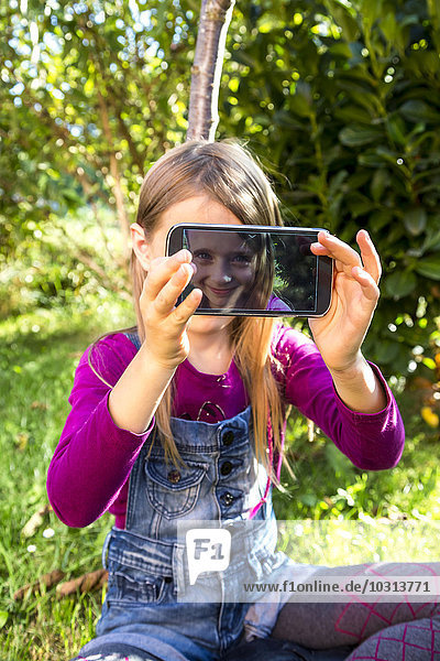 Kleines Mädchen sitzt auf einer Wiese im Garten und zeigt das Display eines Smartphones mit ihrem Selfie.