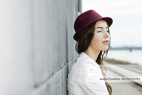 Porträt einer jungen Frau mit geschlossenen Augen  die einen Hut trägt  der an eine Wand gelehnt ist.