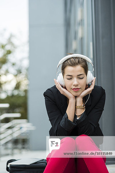Junge Frau mit geschlossenen Augen sitzt auf einer Treppe und hört Musik über Kopfhörer.