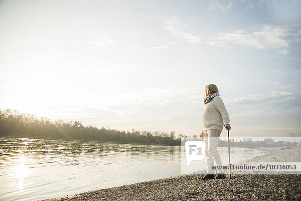 Senior woman with walking stick standing at waterside watching sunset