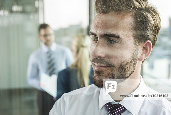 Smiling young businessman looking out of window with colleagues in background