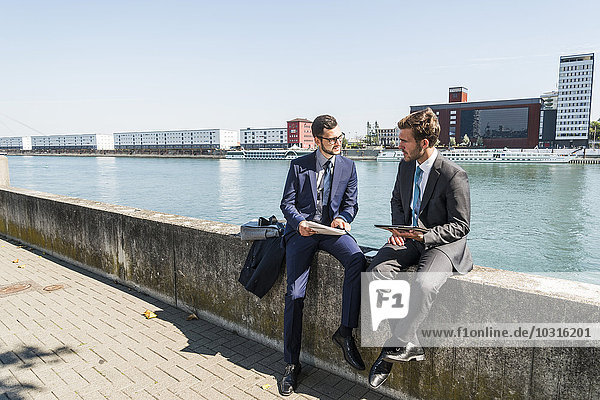 Two young businessmen sitting on wall by river  working