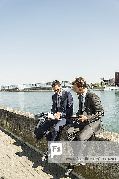 Two young businessmen sitting on wall by river  working