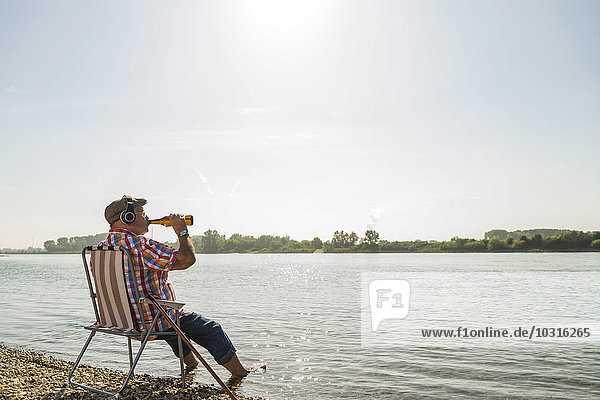 Germany  Ludwigshafen  senior man with headphones sitting on folding chair at riverside drinking beer