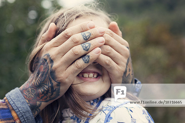 Man's tattooed hands covering eyes of his daughter