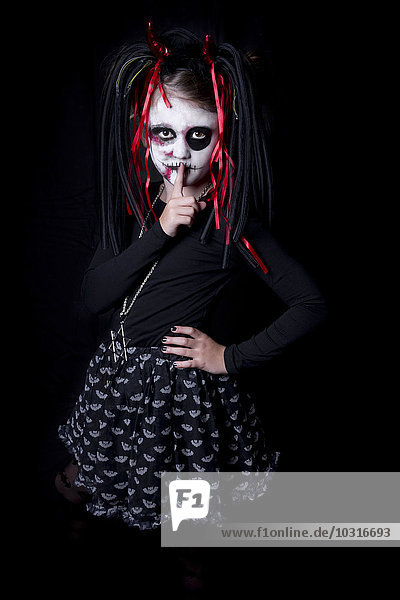 Mädchen mit Finger auf Mund maskiert als Teufel vor schwarzem Hintergrund