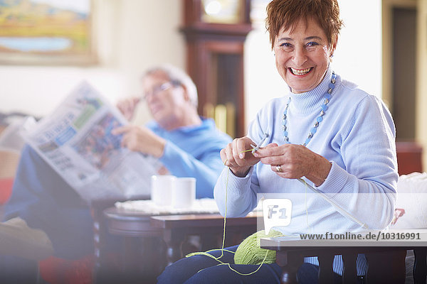 Senior woman knitting with husband in background reading newspaper