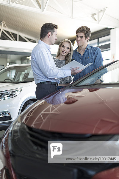 Autohändler zeigt dem jungen Paar eine Broschüre im Showroom