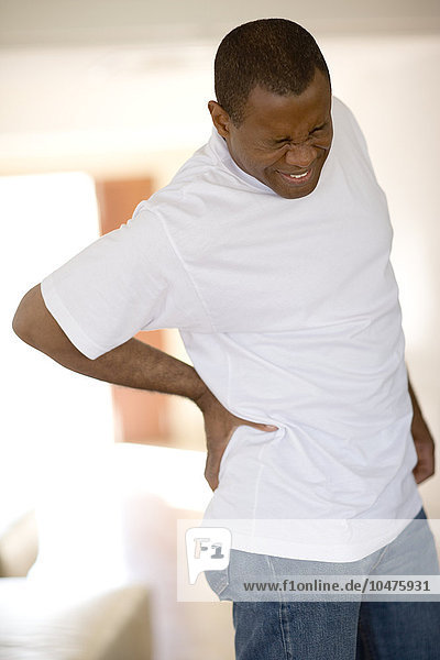 MODELL FREIGEGEBEN. Schmerzen im unteren Rücken. Ein Mann hält sich seinen schmerzenden unteren Rücken. Bei den Schmerzen im unteren Rücken könnte es sich um einen Hexenschuss handeln  bei dem die Weichteile um die Knochen der Wirbelsäule überlastet sind. Dies kann durch unsachgemäßes Heben schwerer Lasten verursacht werden. Die Behandlung umfasst Ruhe und schmerzstillende Medikamente. Schmerzen im unteren Rücken