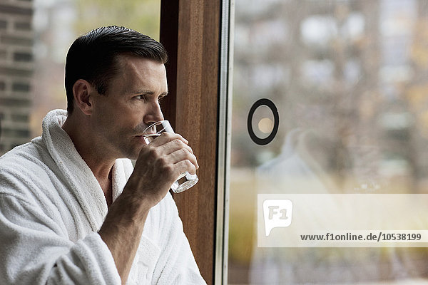 Mann im Bademantel Trinkwasser am Fenster