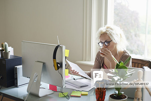 Junge Frau trinkt Tee und liest Papierkram am Schreibtisch im Home-Office.