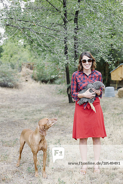 Portrait of a smiling woman holding a grey hen  a dog standing beside her.