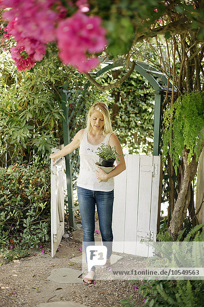 Eine blonde Frau betritt einen Garten durch ein weißes Holztor und trägt einen Pflanztopf.