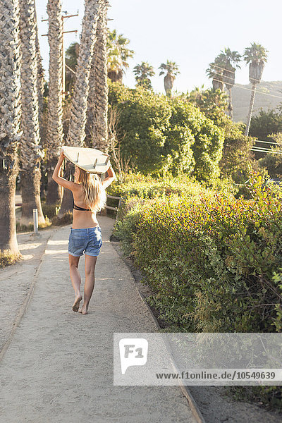 Blond woman in a black bikini and denim shorts carrying a surfboard on a path lined with palm trees.