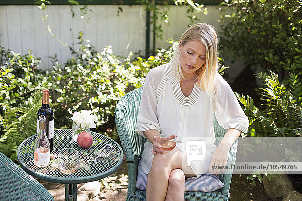 Blonde Frau  die im Sommer in einem Garten sitzt und ein Weinglas hält.