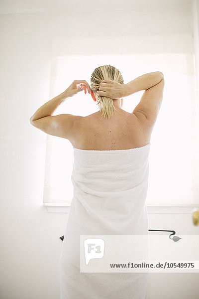 In ein weißes Handtuch gewickelte Frau steht in einem Badezimmer und bindet sich das nasse Haar.