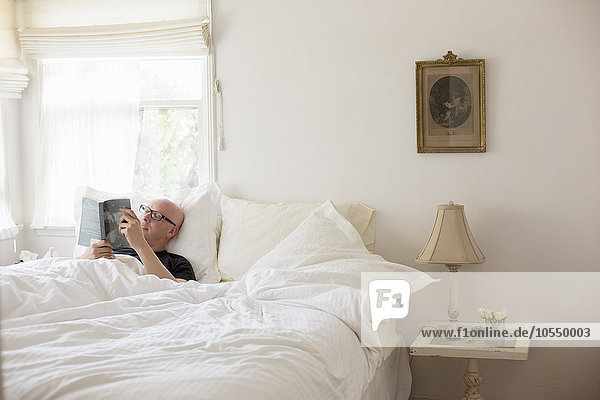 Mann liegt in einem Bett mit weißer Bettwäsche und liest.