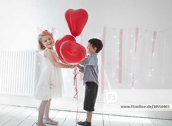 Young boy and girl posing for a picture in a photographers studio  holding red balloons.