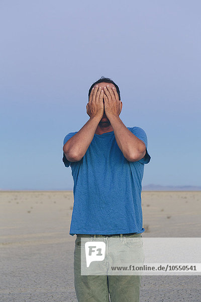 Ein Mann in offener Wüstenlandschaft  der sein Gesicht mit den Händen bedeckt.