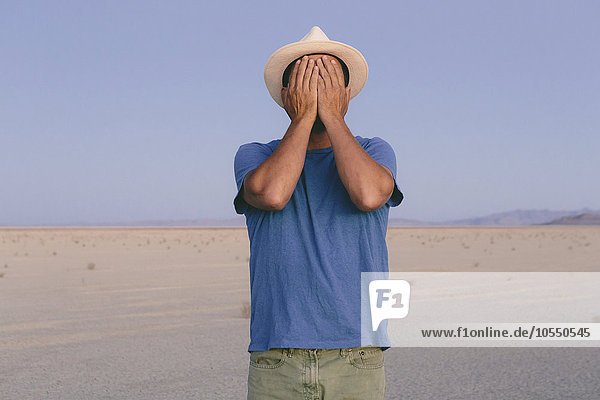 Ein Mann in einer offenen Wüstenlandschaft  der sein Gesicht mit den Händen bedeckt.