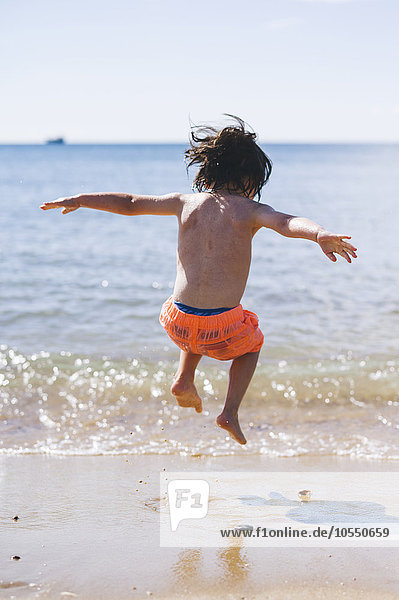 Ein Junge in Badehose  der am Meeresufer über Wellen springt.