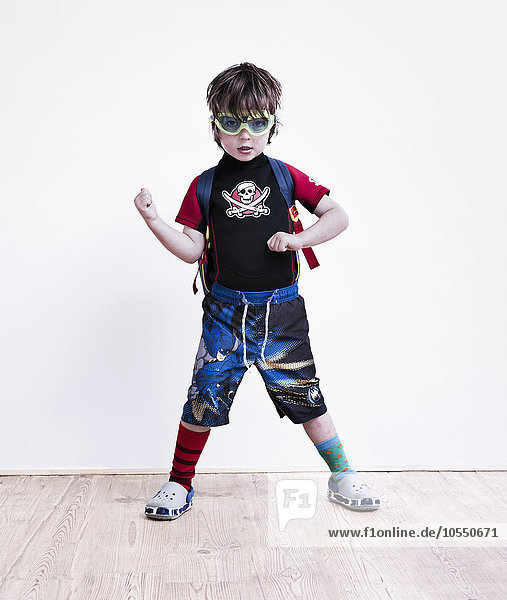Ein Junge steht mit gespreizten Beinen  posiert in einem Kostüm  trägt ein Piraten-T-Shirt  eine Schutzbrille und lange Shorts.