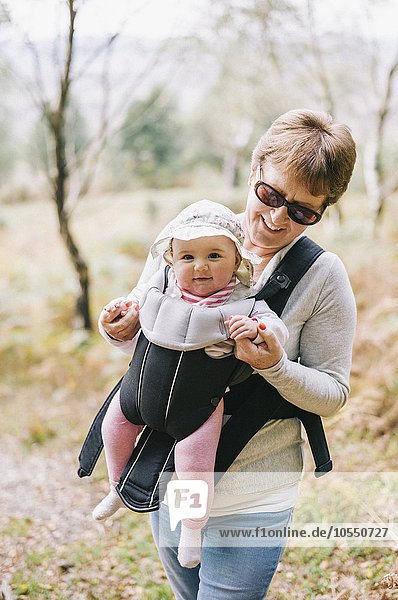 Eine Frau mit Sonnenbrille  die ein Baby trägt  das einen weißen Sonnenhut in einem vorderen Babytragetuch trägt.