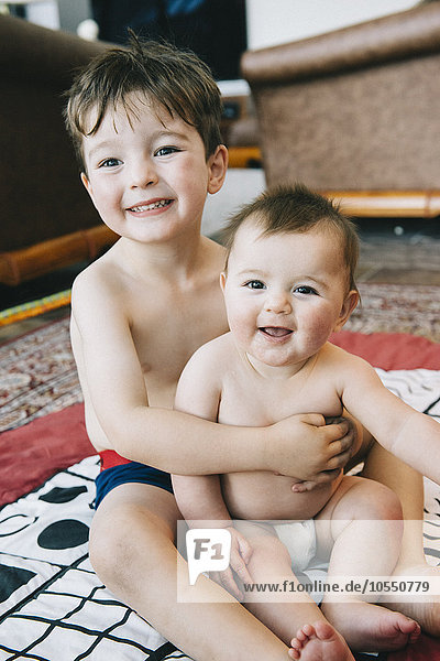 Zwei Geschwister,  ein Junge und seine kleine Schwester sitzen lachend auf einem Teppich.