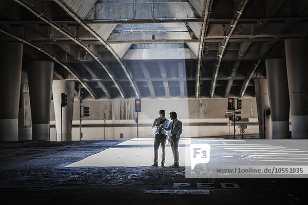 Zwei Männer in einer städtischen Unterführung  die von Beton- und Glaskonstruktionen umgeben sind  nehmen ein Selfie.